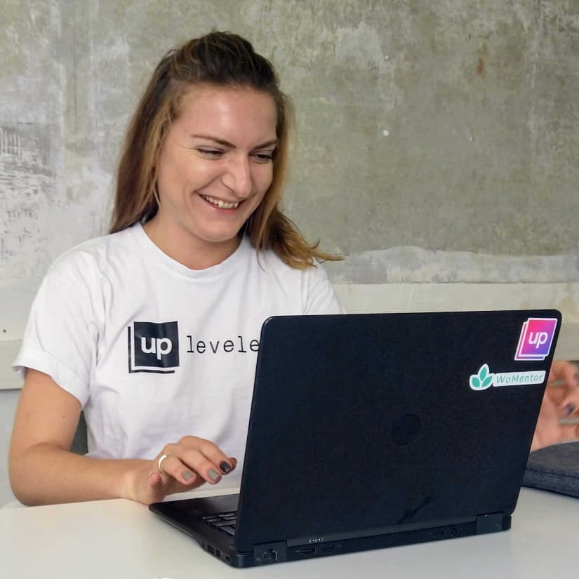 Blonde woman is laughing while working on laptop, wearing a white shirt with an UpLeveled logo print