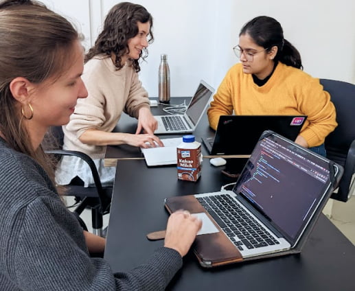 Three young women sit at one desk with their laptops, collaborating on a coding project