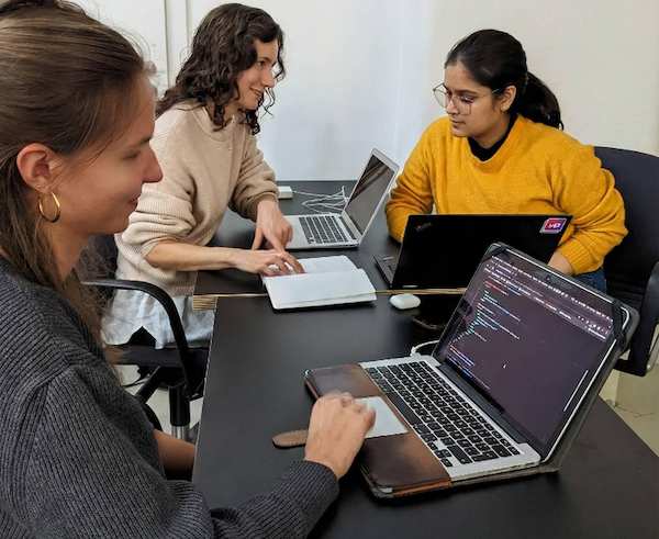 Three young women sit at one desk with their laptops, collaborating on a coding project