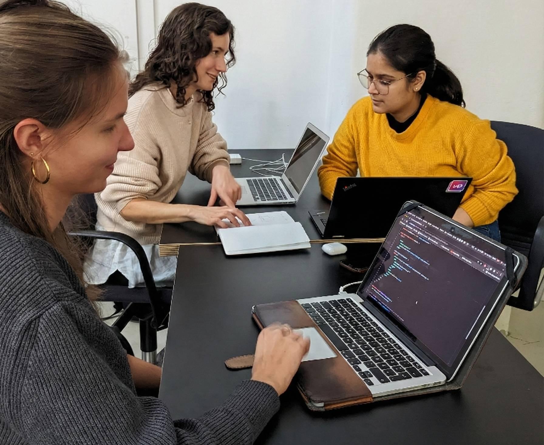 Three young women sit at one desk with their laptops, collaborating on a coding project