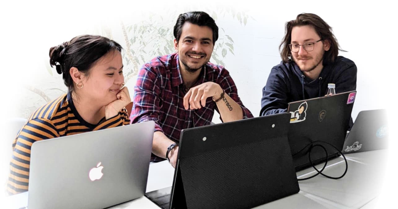 Two male and a female student sit next to each other behind their laptops, smiling