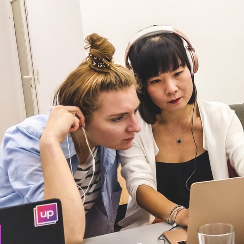 Woman with Asian features working on a laptop, white woman observing