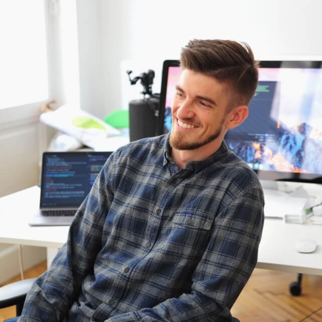Young white man with brown hair and short beard, leaning back in chair smiling, work setup with screens and code in the background