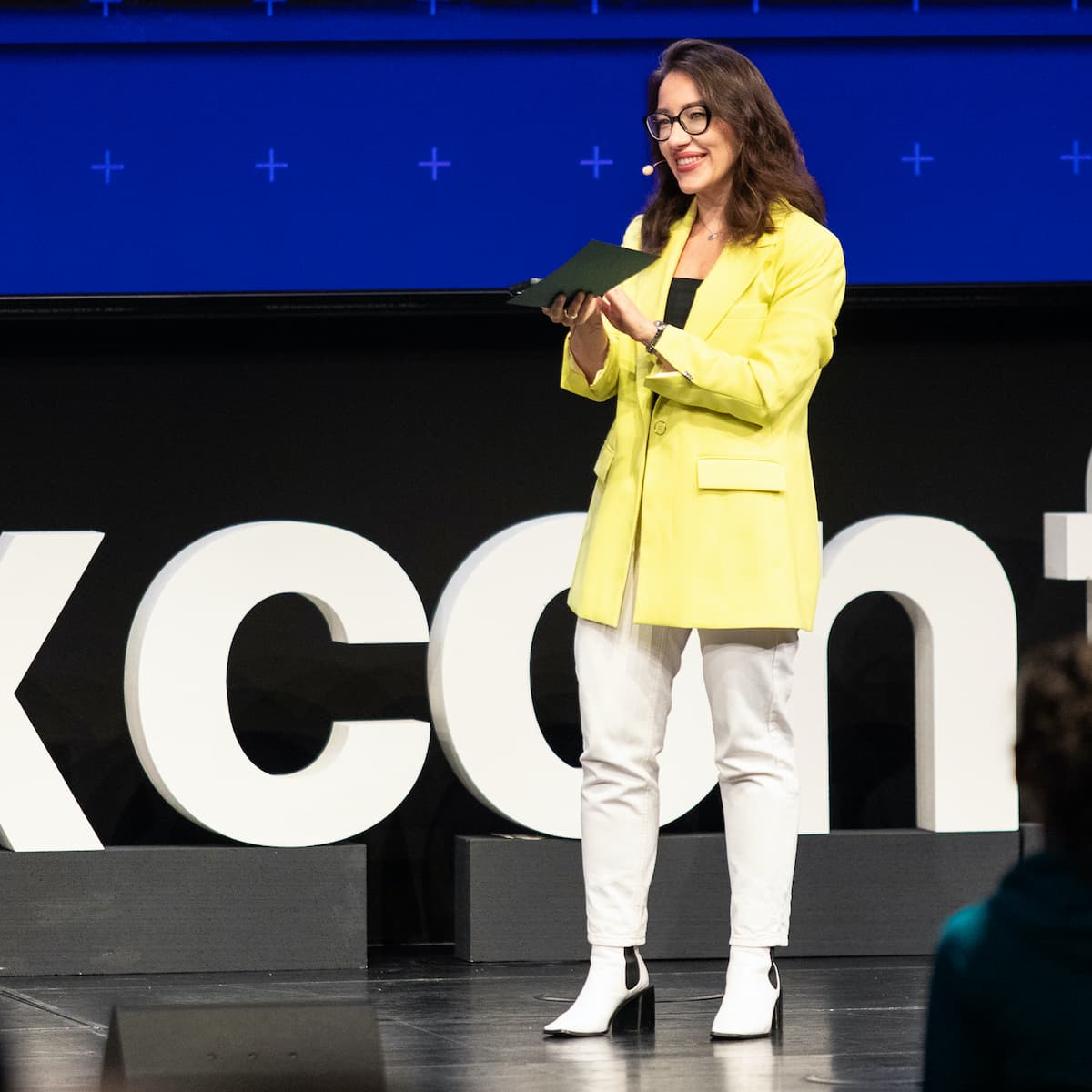 Young woman with long brown hair and glasses speaking on stage of ux conference