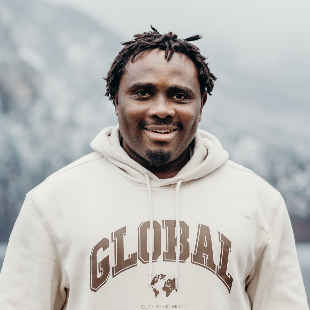 Black man with short dreads in hooded sweatshirt smiles in front of alpine mountain
